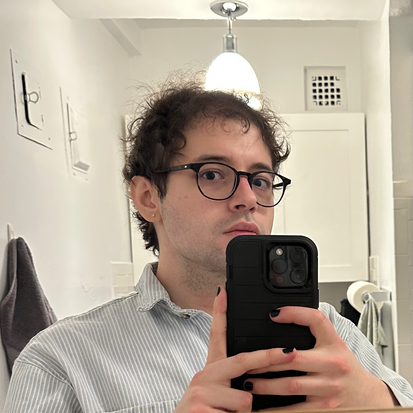 A white woman, wearing glasses, a button-down shirt, and black nail polish, holds her phone up to a mirror and looks into the camera.